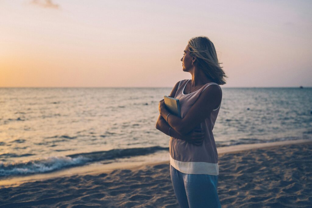 A woman holding a notebook stands on the beach, gazing at a serene sunset.