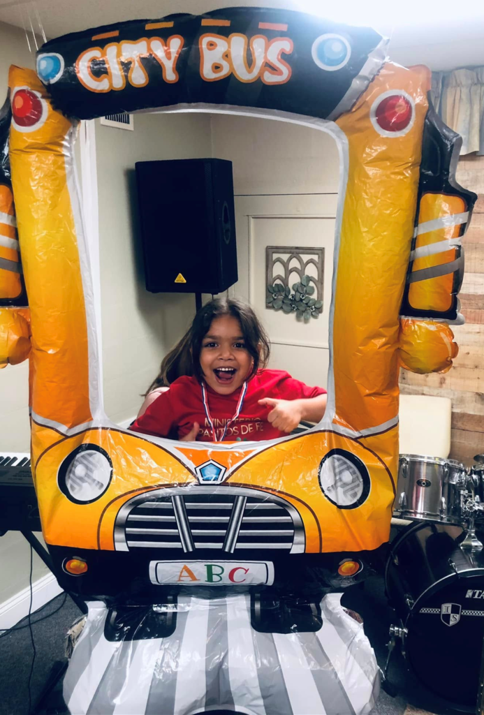 Una niña sonriente se encuentra detrás de una colorida escenografía de “City Bus” usada durante la Escuela Bíblica de Vacaciones. La escena refleja el compromiso del ministerio de enseñar a los niños sobre Dios de manera divertida, significativa y dinámica.