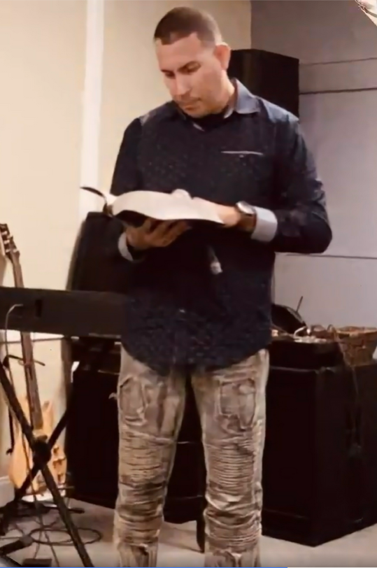 Leslie González lee de una Biblia abierta desde el altar, compartiendo una breve reflexión con los hombres. Su presencia transmite liderazgo, sabiduría y compromiso con el crecimiento espiritual masculino