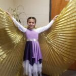 Una niña del ministerio infantil extiende sus alas doradas en una danza de adoración, vestida como ángel y expresando alabanza con gozo a través del movimiento.