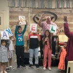 Niños del ministerio sostienen letras coloridas que forman el nombre del pastor, mientras él sonríe detrás del púlpito, rodeado de amor y celebración.