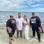Los pastores y la pareja Kimberly y Jesús Manuel están juntos en la playa después de su bautismo, sonriendo con gozo mientras celebran este paso sagrado de fe.