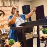 La pastora Wanda está detrás del púlpito con el micrófono en una mano y la otra levantada en alabanza, iniciando el servicio con pasión y reverencia.