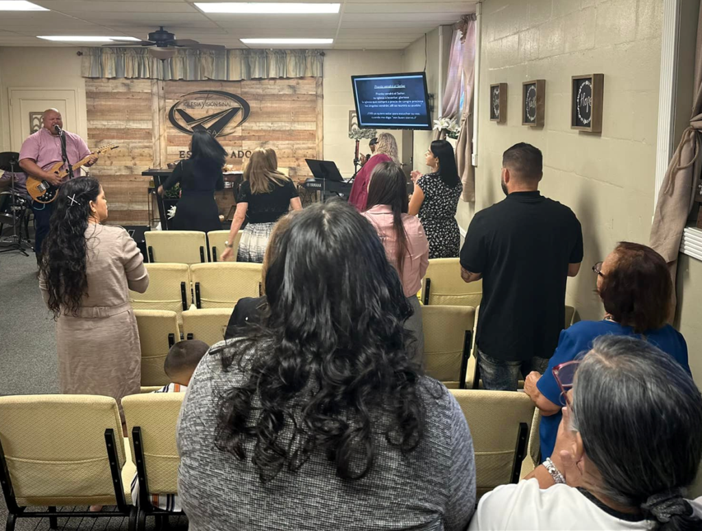 Vista desde el fondo del santuario donde la congregación adora con pasión mientras el pastor dirige con su guitarra desde el altar, reflejando el ambiente lleno del Espíritu y la alegría del servicio dominical en Vision Sinai.